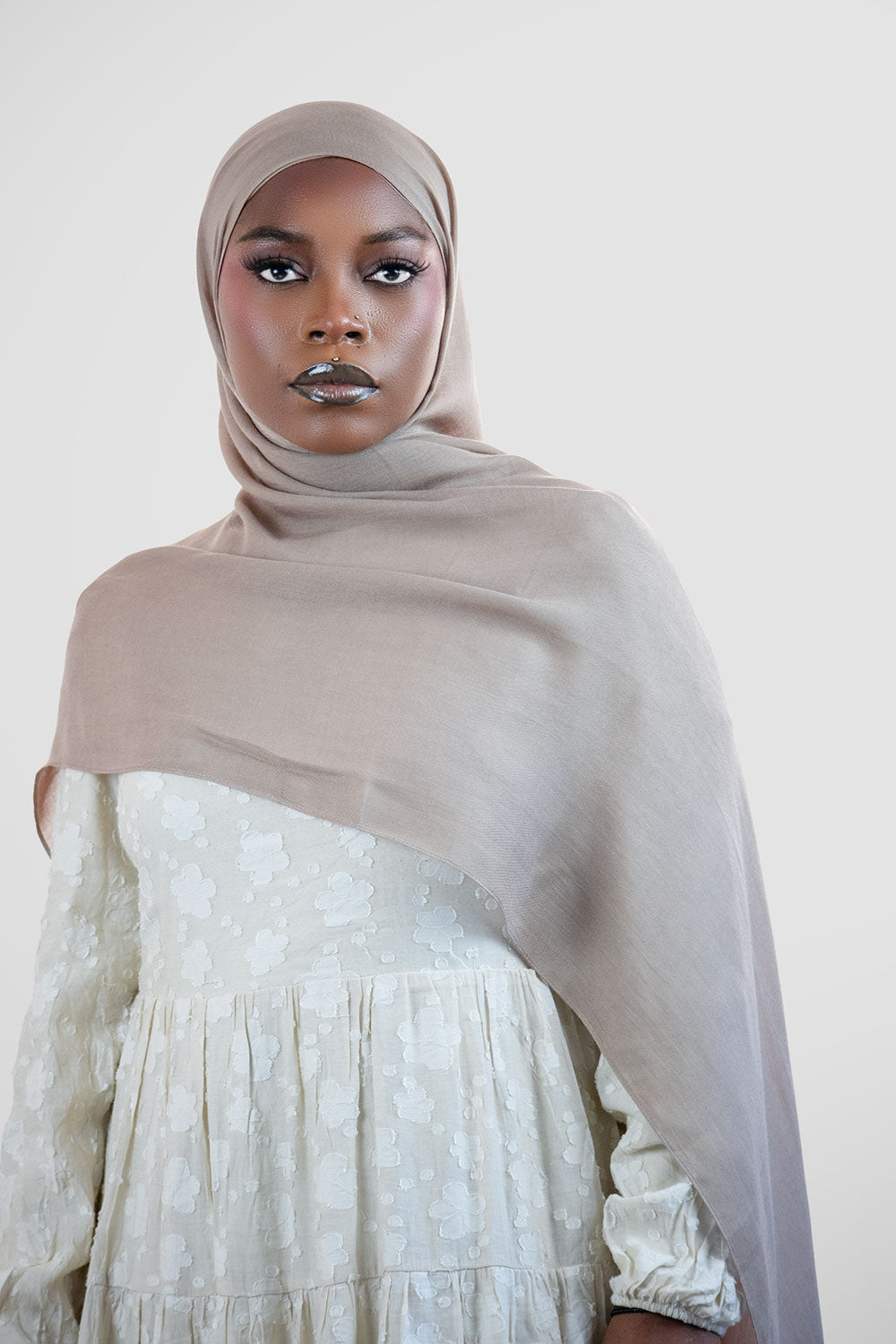 Woman wearing a gry modal  hijab and white dress against a plain background