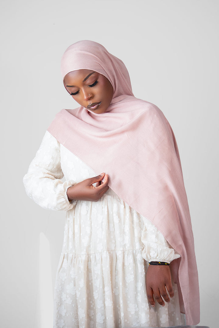 Woman wearing a light pink hijab and white dress against a plain background