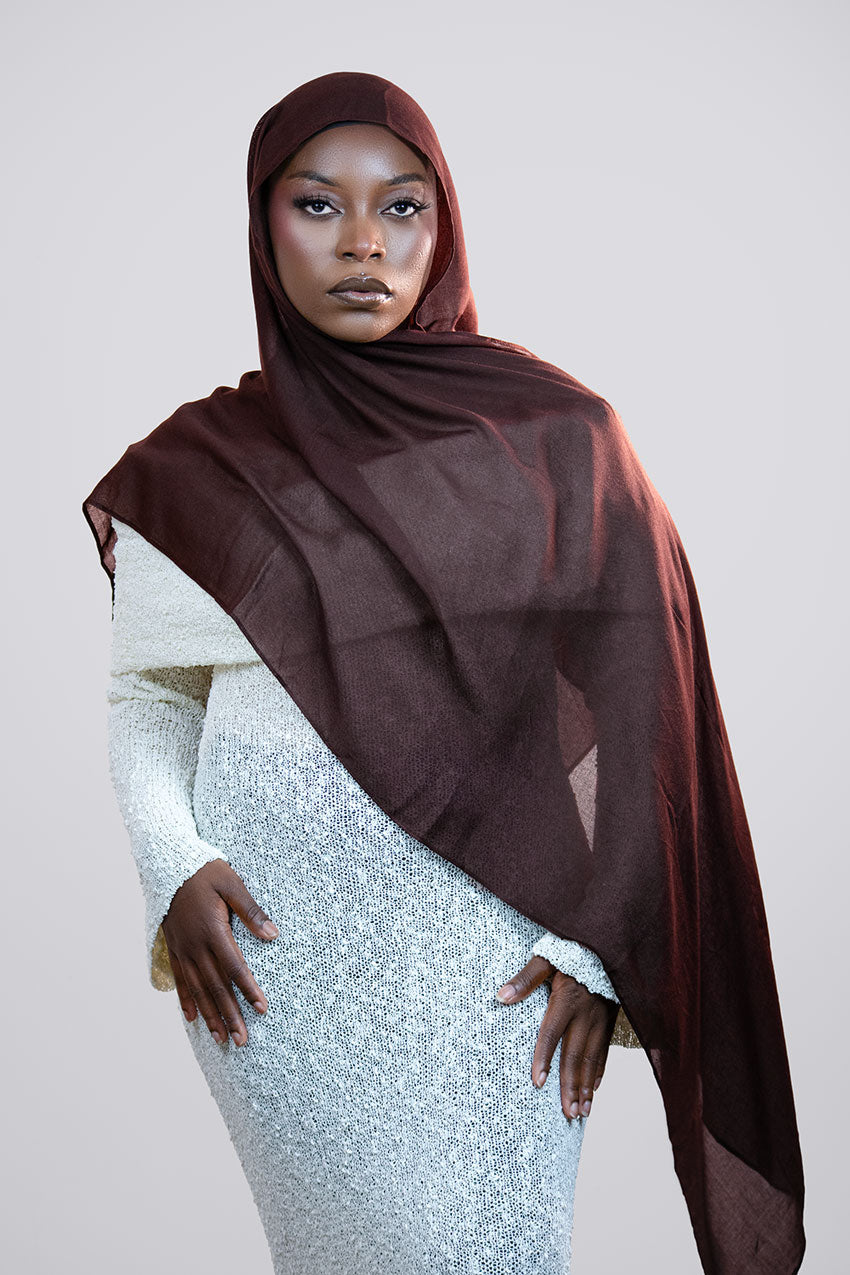 Woman wearing a brown hijab and a white dress against a plain background
