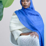 Woman wearing a blue hijab and white outfit sitting on a chair with a plant in the background