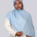 Woman wearing a light blue hijab and beige dress against a plain background