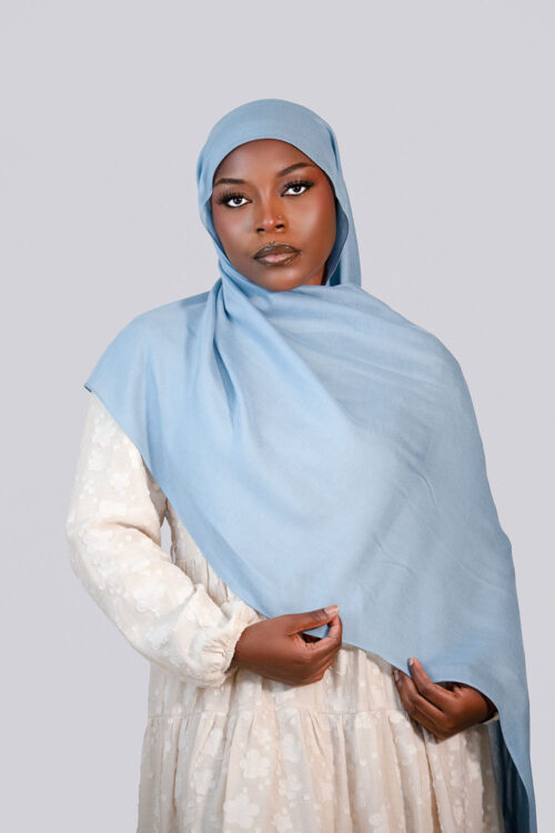 Woman wearing a light blue hijab and beige dress against a plain background